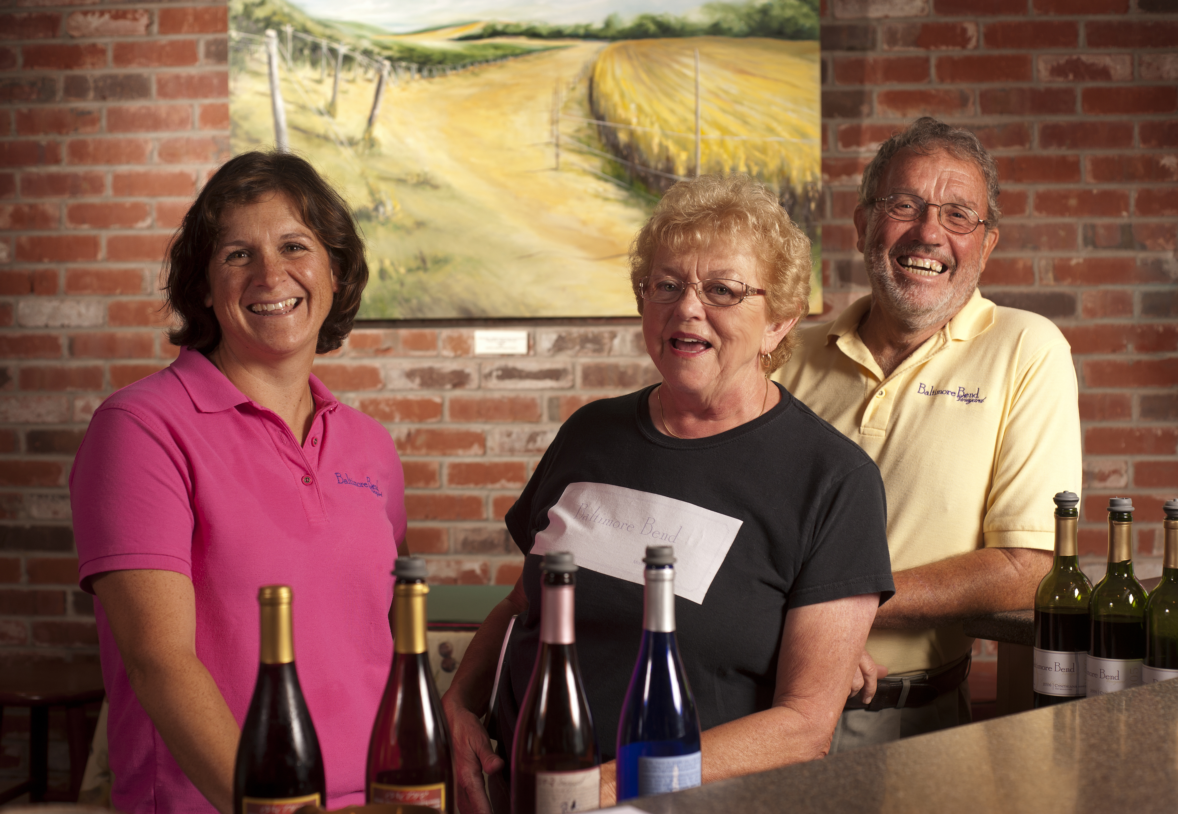 Baltimore Bend Vineyard - indoor photo, three people smiling while standing in front of a bar with wine bottles on it.