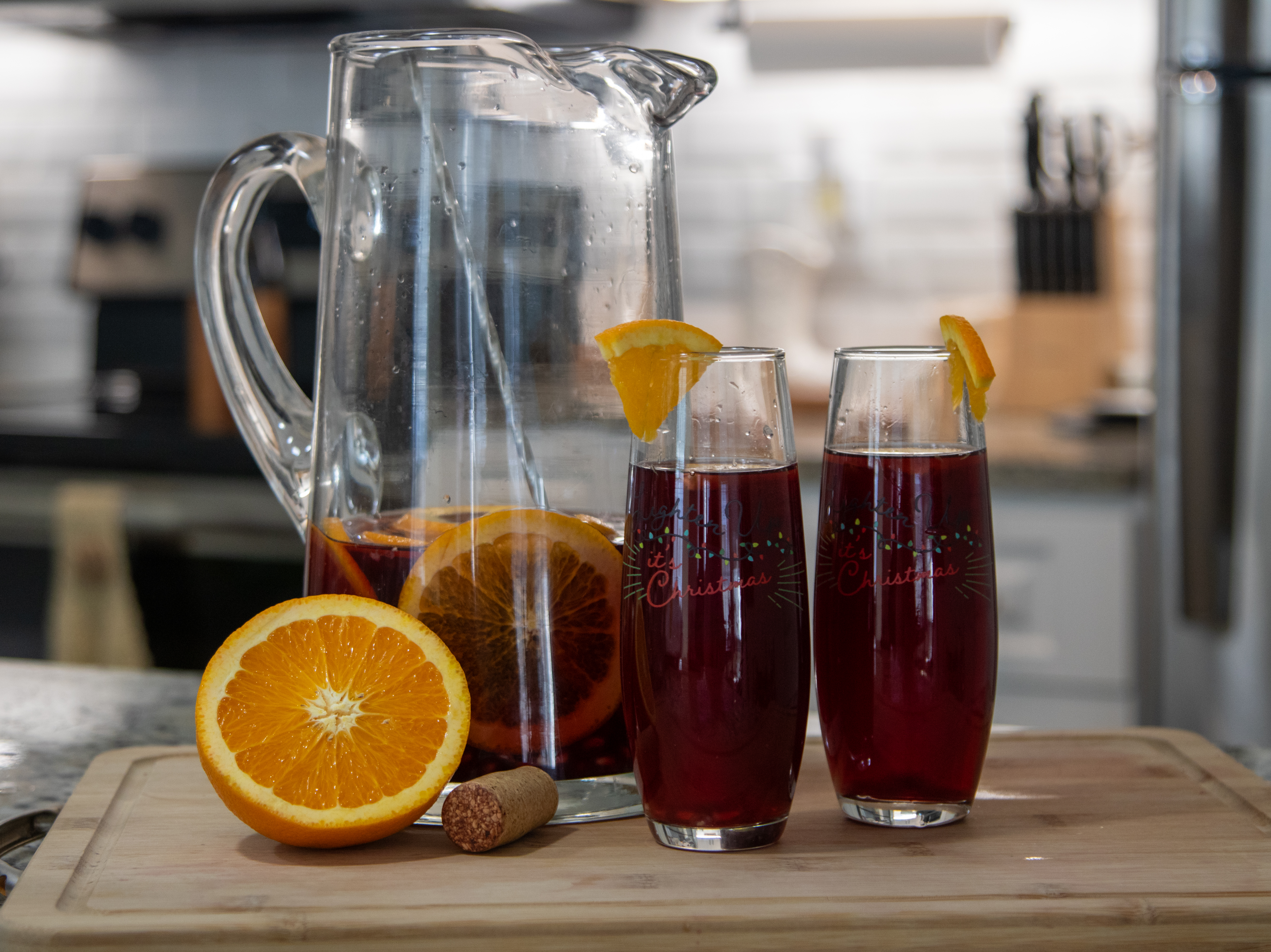 Glass pitcher half full of sangria. 2 glasses of sangria with orange slices on the rims. There is a half cut orange with a cork sitting in front of the pitcher.